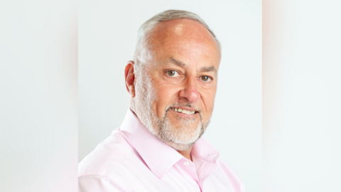 Portrait photo of councillor Mark Howard. He is wearing a pink shirt and has combed, white hair and a white beard. He is smiling.