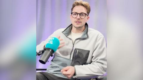 A man wearing glasses in a zip top, speaks to the camera while sitting behind a blue mic