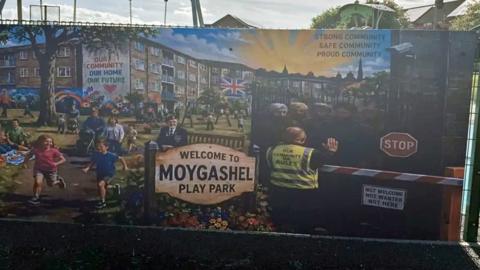 Banner on a play park fence, showing children playing sunshine and men being rejected at a barrier.