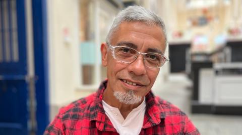 A brown skinned man with grey hair and a goatee beard looking towards the camera. He's wearing glasses and a red and black checked shirt. 