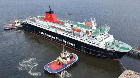 A black and white ship with red funnels leaving a dry dock. Two tugs at the side of the ship and rear are helping it leave the dock