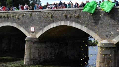 People on the bridge emptying bags of ducks. There are two people emptying the bags while other people along the bridge watch on. The bridge has three arches and there is a river below.