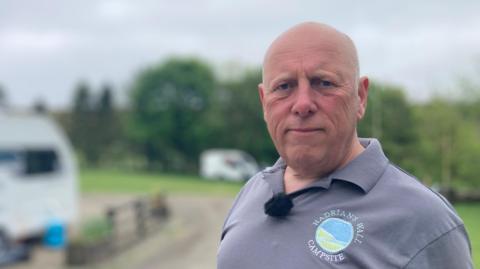 Steve Miller is standing on his campsite and looking at the camera. He is bald and is wearing a grey polo shirt with a "Hadrians Wall Campsite" logo on it.