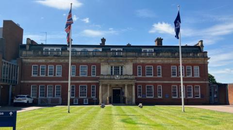 A large brick building with bay windows. A lawn is in front of the building with two flags - the Union Jack and Northamptonshire Police logo - flying.