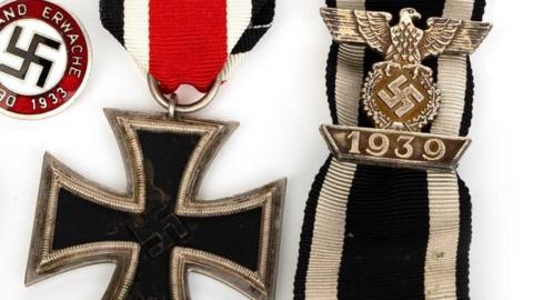 Nazi-era German military medals, including a black Iron Cross and German Nazi eagle lie next to a swastika badge on a while background. 