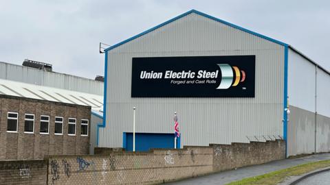 The Union Electric Steel building which is a grey, corrugated building at the side of a side. It has a black sign in the centre which reads 'Union Electric Steel forged and cast rolls.'