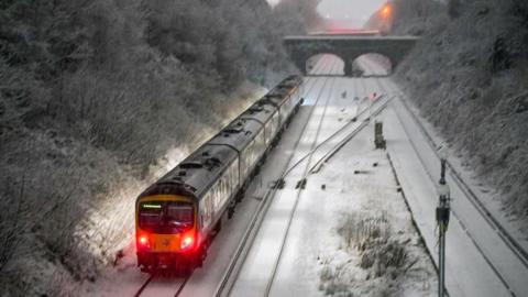 A Merseytrail train operating in snowy conditions, with its lights on at the front and a sign saying 'Liverpool'. Trees and shrubs either side of the line have a covering of snow and the glow of an amber street light can be seen on a bridge behind the train.