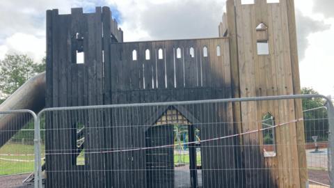 A large wooden castle-like structure is severely burnt with char marks across most of the surface. It is surrounded by a large metal fence.