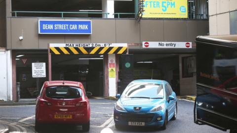 A red and blue car and the edge of a black van parked in front of a multi-storey car park building. It is a large brown building with Dean Street Car Park written inside a blue box. There is a white sign above the entrance which reads: 'Max height 2.0m- 6'7". There is a no entry sign above the exit on the right.