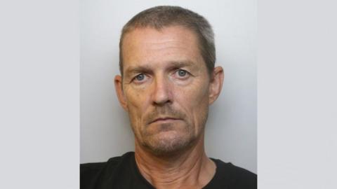 A police mugshot of Nigel Frith. He is wearing a black t-shirt and standing against a grey background. He has short brown hair which is turning grey and a closely shaven goatee style beard.