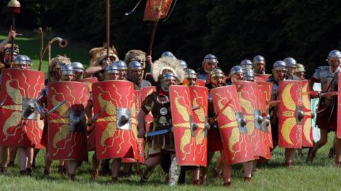 Modern Roman military reenactors, holding large rectangular red shields and wearing polished metal armour, form a line as if preparing for battle