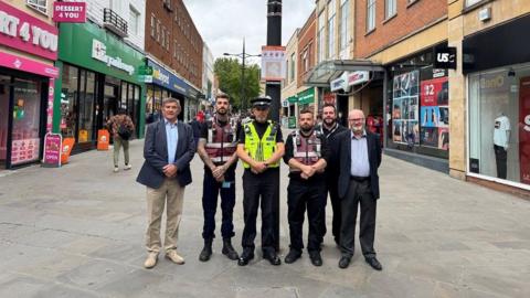 A group of six men in uniform, including a police officer, community support officers and people in suits, stand in the middle of a pedestrian area in Swindon town centre. Shops are either side of them.