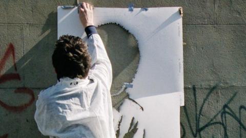 A picture taken from behind showing a man wearing white overalls, holding up a stencil onto a grey brick wall.