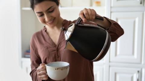 A young woman pouring water from a kettle into a cup. 