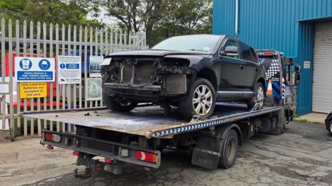 A damaged large black SUV style car on the back of a recovery vehicle. The vehicle is parked in what looks like an industrial estate.