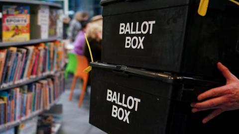 Two black boxes with Ballot Box written on the front are being carried past a bookshelf packed with books.