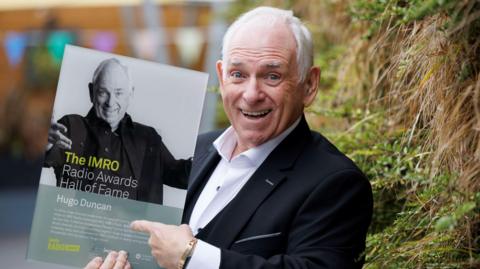 Hugo Duncan smiling widely, holding a picture of himself on which are written the words IMRO AWARDS HALL OF FAME HUGO DUNCAN