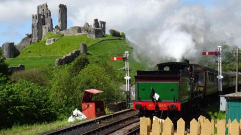 A steam train - painted red, green and black - on the railway line by Corfe Castle ruins, which are mounted on a grassy hill. There is steam billowing from the train.