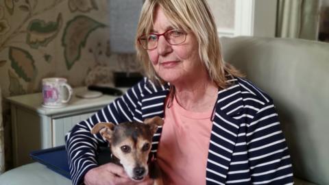 A woman with blonde shoulder-length hair and wearing glasses. She is sitting in an armchair, with her Jack Russell dog on her lap. She is wearing a navy and white striped jacket with a coral coloured T-shirt.
