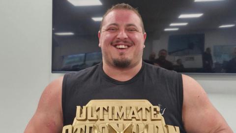 Dean Mason - a man with broad shoulders and muscles - has short brown hair and stubble and is smiling into the camera. He is wearing a black vest and holding a gold trophy that says ultimate strongman.