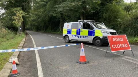 A country road which goes through Rivelin Valley in Sheffield. The photo is taken from just behind some police tape. A sign in line with the tape reads "road closed". Just behind the sign is a police van with the front doors open.