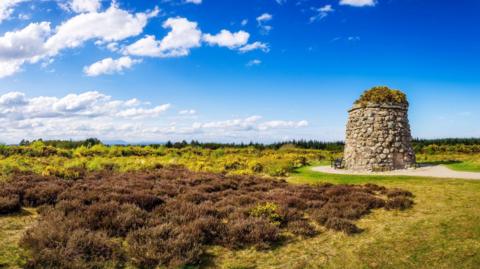The memorial is a tall, stone-built structure with vegetation on top. It is in an area of sweeping moorland.