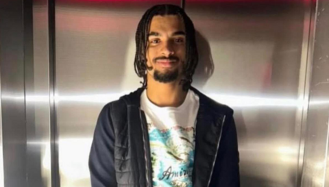 Lain Estridge, who has dreadlocks and a goatee beard. He is wearing a white t-shirt with a tropical image on it, and a black puffer-style body warmer. He appears to be standing in front of the doors of a lift. 