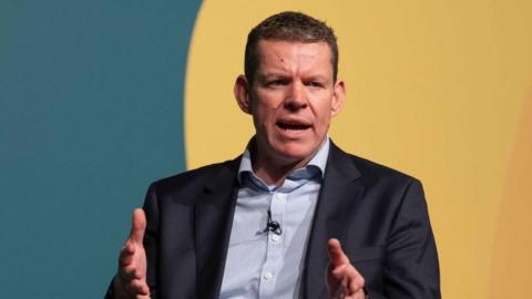Plaid Cymru Leader Rhun ap Iorwerth speaking at a conference. He is sitting in front of a green and yellow wall. 