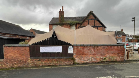 The brown fabric of the tented marquee spans an outdoor area at the back or side of the pub. It is behind a medium-sized wall with the pub then behind the marquee. A car park can be seen on the right of the image.