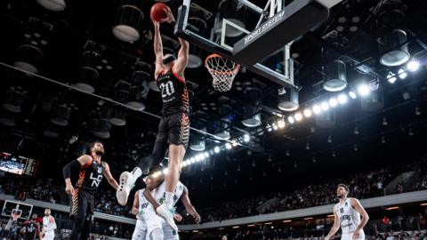 London Lions basketball player in black jersey jumps into the air holding the basket ball close to the hoop in an arena. There are players wearing white kit nearby and there is a crowd in the background.