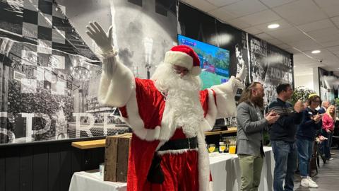 A man dressed as Father Christmas stands with his arms open wide and there are people around him clapping