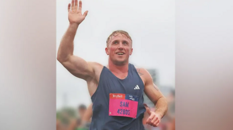 A runner wearing a vest with the words Sam on the front, smiling and with one hand in the air 