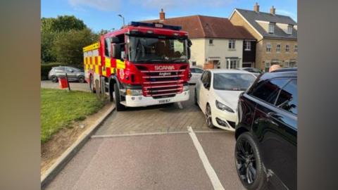 A fire engine is trying to turn around a corner, but has had to go over a grass verge due to the parking of a car on the other side of the road.