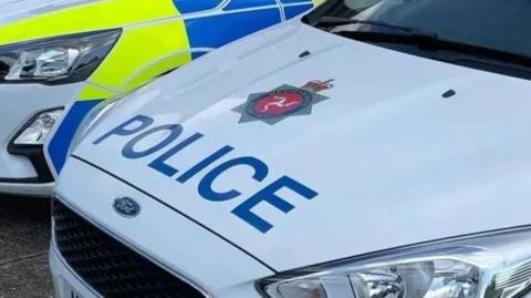 The bonnet of a white car which reads police in blue and has the three legs of man in a star which is the police logo.