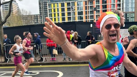 A man running a race is wearing a rainbow coloured outfit and hat, along with sunglasses. He is waving to the camera.