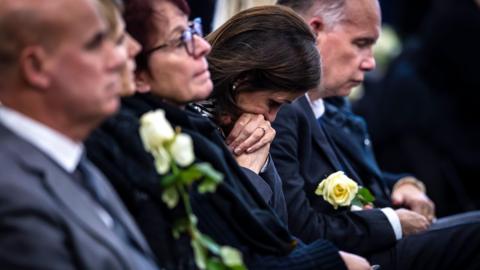 A woman rests her face on her hands. She sits in a row of people wearing dark colours, some hold white roses