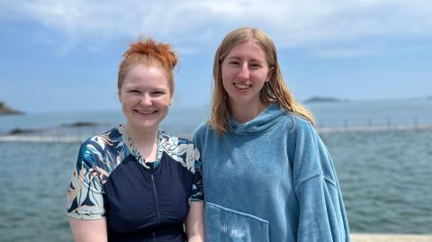 Two young women looking at the camera smiling, stood in front of the sea.