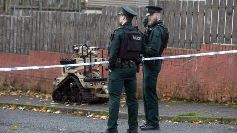 Two male police officers standing at a blue and white police tape. Their faces are blurred and they are wearing a bottle green police uniform with a black bullet proof vest. There is an ATO machine which is a brown colour with black wheels.