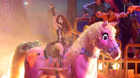 Chappell Roan singing wearing a glittery outfit on a large pink pony prop on stage