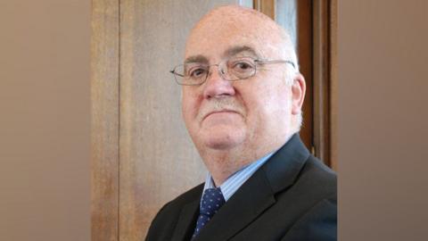 Chris Blakeley has short grey hair and a grey moustache and is wearing metal framed glasses. He is wearing a light blue shirt, a navy blue tie dotted tie and a blue suit  jacket.