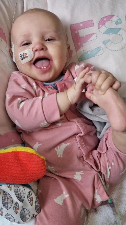 Melody at a few months old is lying in a cot with her mouth open happily looking at the camera. She has hold of her foot and wears a pink babygrow with white rabbits printed on it. She has a tube with a plaster sticking it to her face.