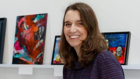 Phil Batty pictured wearing a red and blue striped top. She is sitting to the right of a white wall which includes a shelf with her framed artwork on. She has brown hair.