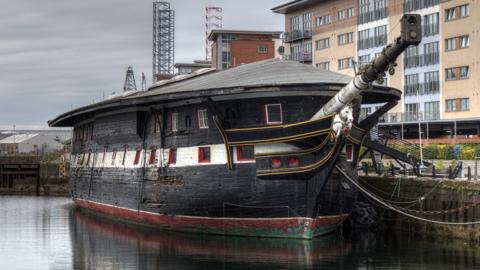 A large black ship sits in a dock with a building in the background