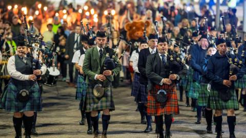 A row of pipers lead the torchlight procession through Fort William. The pipers are wearing different tartans and behind them a large number of people carry flaming torches.