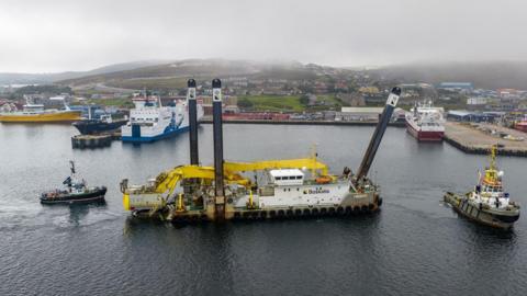 An aerial view of Magnor. The dredger is a long, white vessel carrying yellow excavators. The ship has three long steel piles, which are raised up from the sea. Two tugs are next to the dredger. Moored in the harbour are a ferry and a large fishing boat.