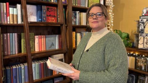 A woman with brown hair tied back is wearing glasses, a white jumper and a green cardigan. She is holding a white book called "Rebecca". Behind her are bookshelves stacked with books, and a wooden ladder leaning against them, with fairy lights nearby.