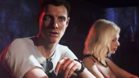 A screengrab from the new Bond video game. Two people are sitting close together in what looks like a warm, low‑lit bar or club. The lighting is mostly red and blue.. The person in the foreground (a young James Bond) is wearing a light shirt with sunglasses hanging from the collar, plus a wristwatch. They’re leaning slightly forward, relaxed, with their arm resting on the top of a bar. Just behind them, a woman with light, shoulder‑length hair is wearing a sleeveless top and a necklace. She's turned slightly away from the camera, and the lighting catches their face more softly.