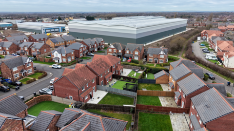 An aerial view of homes next to the under construction Astley Business Park warehouses on December 30, 2025 in Wigan, England. The Astley Warehouse Action Group, which is opposed to the warehouse project, have argued that construction of the large warehouses behind the Henfold Estate has caused property damage and breathing difficulties for those nearby. They also object to the aesthetic impact of the buildings.
