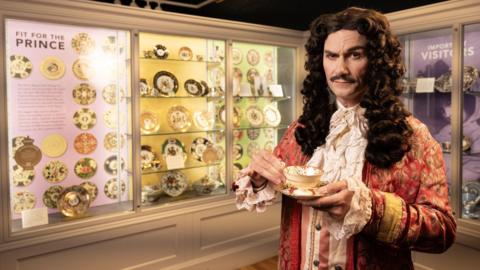 A man wearing a long curly dark brown wig, wearing 17th century clothing which consists of a red and gold coat with white frills on the sleeves, and white frills down his chest and hear his neck. He is holding a china cup and saucer, and is standing in front of a museum display of china plates
