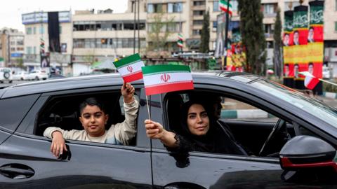 A dark-haired woman and a young boy in a sweatshirt wave small Iranian flags from the windows of a black car. The flag is a horizontal tricolour with green, white and red stripes, and an Islamic central emblem. Photo: 25 March 2026